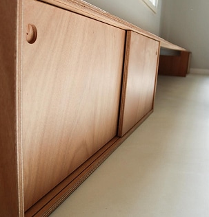 Gros plan sur une commode basse en contreplaqué clair avec portes coulissantes et une poignée circulaire incrustée, vue en angle bas.