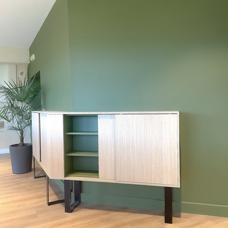 Buffet bas moderne en bois clair avec portes coulissantes, ouvert sur des étagères intérieures vert sauge, contre un mur vert olive.