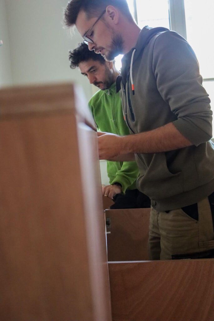 Deux hommes concentrés sur l'assemblage de meubles en bois clair, l'un portant des lunettes et un sweat à capuche vert olive.