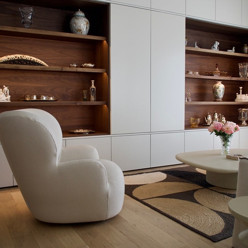 Fauteuil blanc moelleux dans un salon moderne avec étagères intégrées en bois sombre remplies d'objets décoratifs et portes blanches.