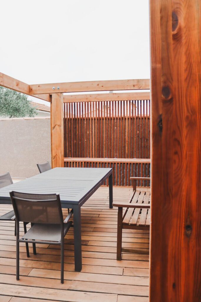 Terrasse en bois sous pergola avec table moderne grise et chaises en textilène, vue à travers un poteau en bois foncé.