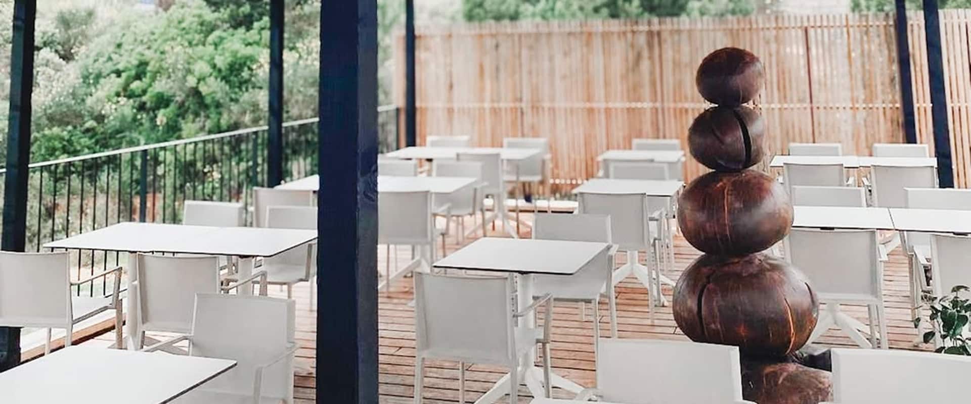 Restaurant extérieur avec tables et chaises blanches sur terrasse bois, devant une cloison en bambou et vue sur verdure.