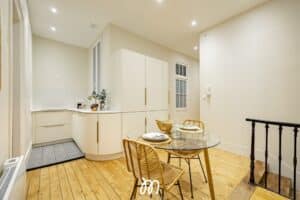 Cuisine-salle à manger moderne claire avec armoires crème, sol en bois clair, table ronde en verre et chaises en rotin. Logo L'Immovation.