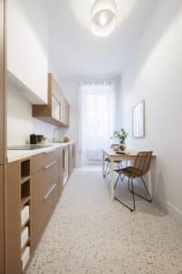 Cuisine étroite moderne avec meubles couleur taupe, plan de travail blanc, sol terrazzo clair, et petite table à manger avec chaises en rotin.