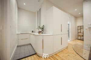 Cuisine moderne crème avec armoires hautes, robinetterie laiton, sol bois clair et tapis géométrique. Logo L'immovation.