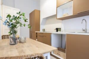 Cuisine moderne avec meubles couleur camel, table en bois clair avec vase de verdure, et porte blanche ouverte.