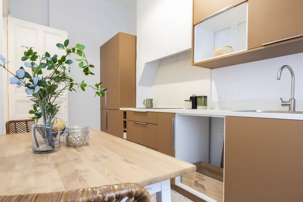 Cuisine moderne camel et bois : design épuré Cuisine moderne avec meubles couleur camel, table en bois clair avec vase de verdure, et porte blanche ouverte.