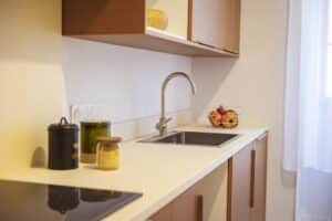 Plan rapproché d'un comptoir de cuisine moderne avec évier en inox, robinet et ustensiles de cuisine, près d'une fenêtre avec voilage.