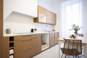 Cuisine moderne avec meubles couleur camel, comptoir blanc, plaque à induction, évier et table à manger en rotin près d'une fenêtre voilée.