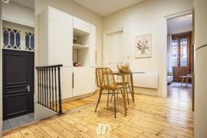 Hall d'entrée lumineux avec sol en parquet clair, rangement blanc, porte d'entrée noire et petit coin repas près d'une affiche 'Flower Market Amsterdam'.