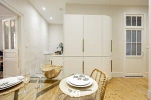 Salle à manger moderne avec table en verre et chaises en rotin, face à des placards blancs épurés et un sol en bois clair. Texte visible : L'IMMNOVATION.